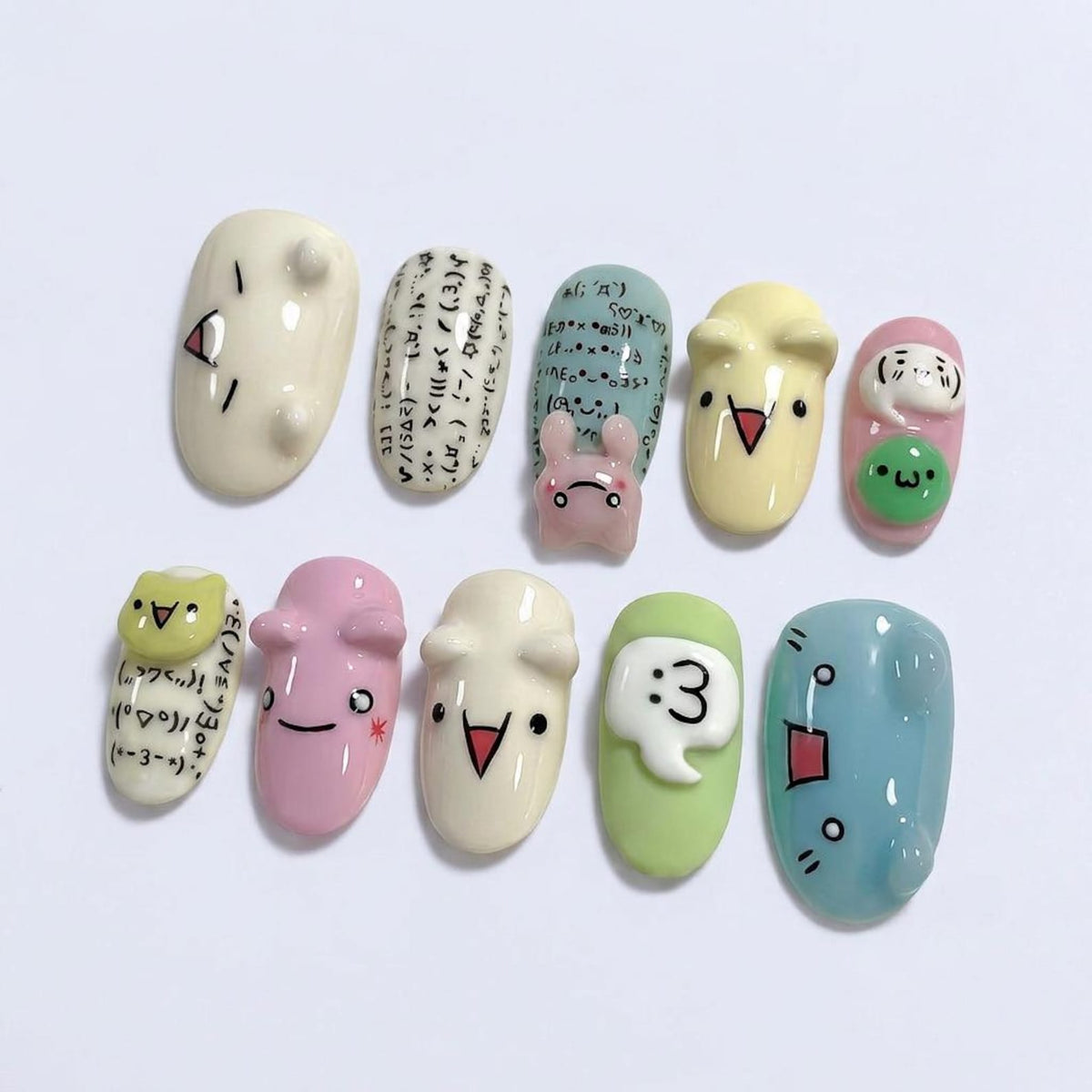 A set of eleven painted artificial nails with various cartoon designs. The nails feature a variety of colors, including white, pink, yellow, green, and blue. Some nails have faces, while others have text and symbols.