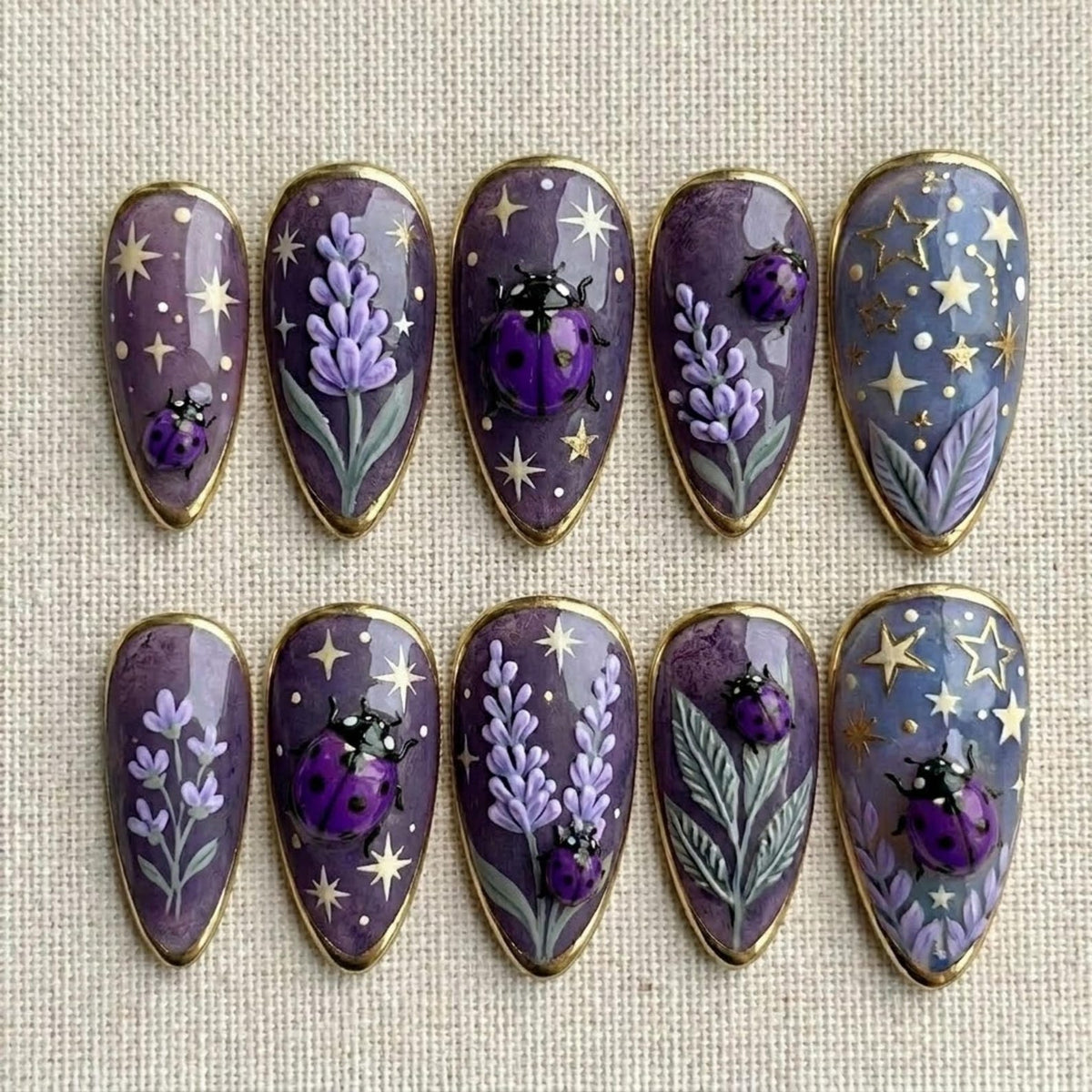 A set of ten almond-shaped, purple nail art designs. Each nail features a gold border and unique designs, including lavender sprigs, ladybugs, and star patterns. The ladybugs are a vibrant purple.