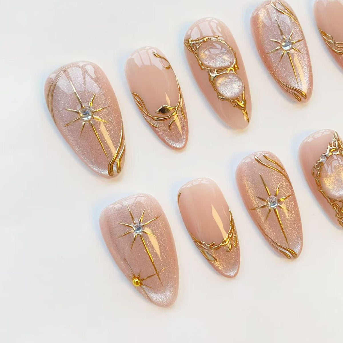 A set of almond-shaped false nails in a light pink hue, adorned with gold metallic accents. The designs include starbursts, swirling lines, and ornate embellishments, creating a glamorous and elegant aesthetic.