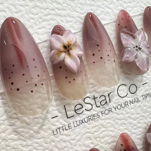 Close-up of several almond-shaped artificial nails. The nails feature a gradient design, transitioning from a light beige to a mauve hue, with scattered dark red dots. Some nails are adorned with 3D p