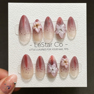 A set of oval-shaped false nails with a gradient design, transitioning from a clear base to a reddish-brown hue. Some nails feature small black dots and 3D flower embellishments. The text "- LeStar Co