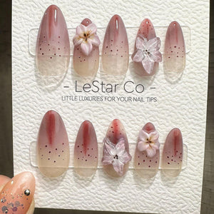 A set of almond-shaped false nails with a gradient design, transitioning from a nude base to a reddish-pink tip. Some nails feature small dark dots and 3D flower embellishments. The text "- LeStar Co 