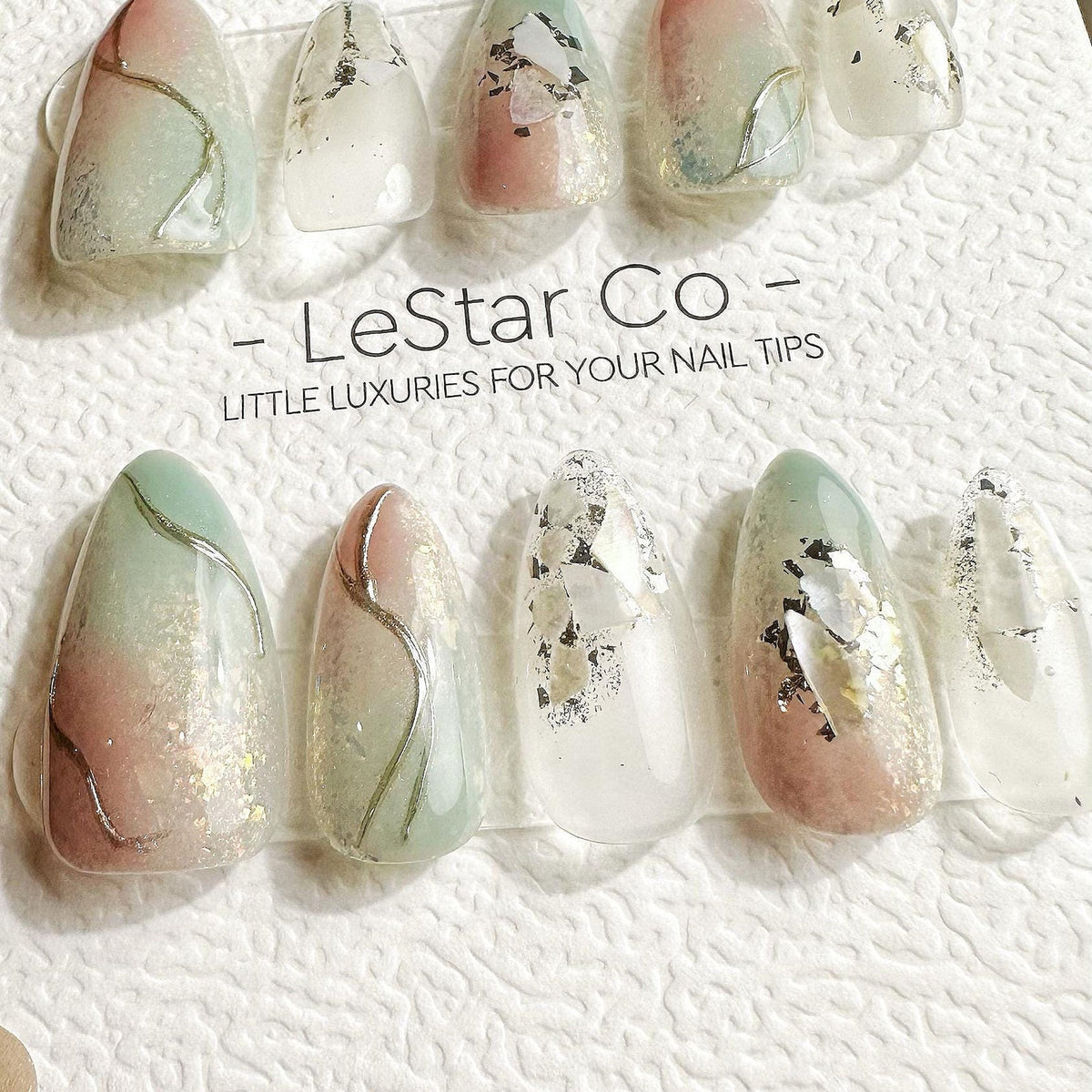 A set of artificial nails with a variety of designs. Some nails feature a gradient of pink and green with silver accents. Others have silver foil or glitter embedded in a clear base. The text "- LeSta