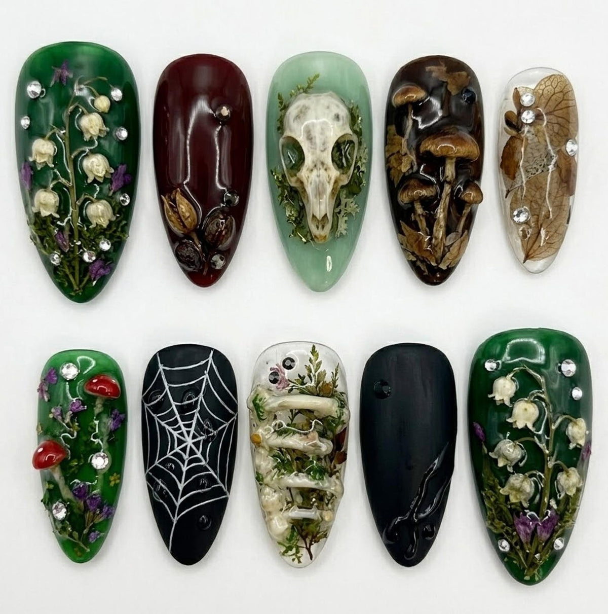 A collection of ten artificial nails with various designs. The nails feature botanical and gothic themes, including mushrooms, a spiderweb, a skull, and floral arrangements. Colors include green, burgundy, black, and clear, with some nails adorned wi