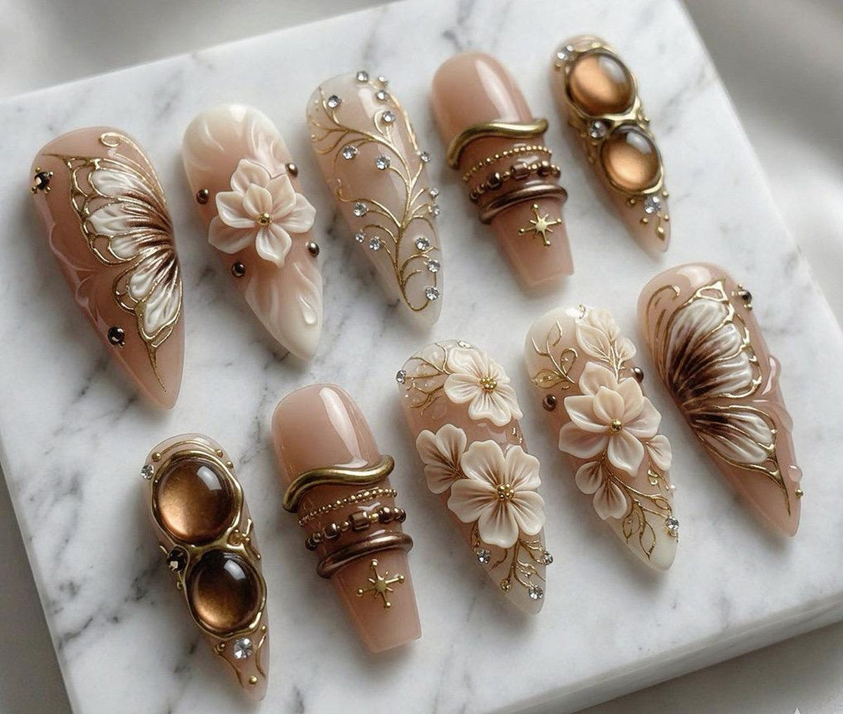 A collection of ten almond-shaped artificial nails. The nails feature various designs, including floral embellishments, gold accents, and jewel-like adornments. Colors include nude, gold, brown, and white. Some nails have a butterfly wing design.