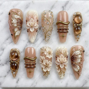 A collection of ten almond-shaped artificial nails. The nails feature various designs, including floral patterns, butterfly motifs, and gold embellishments with rhinestones. The color palette includes shades of beige, brown, and gold.