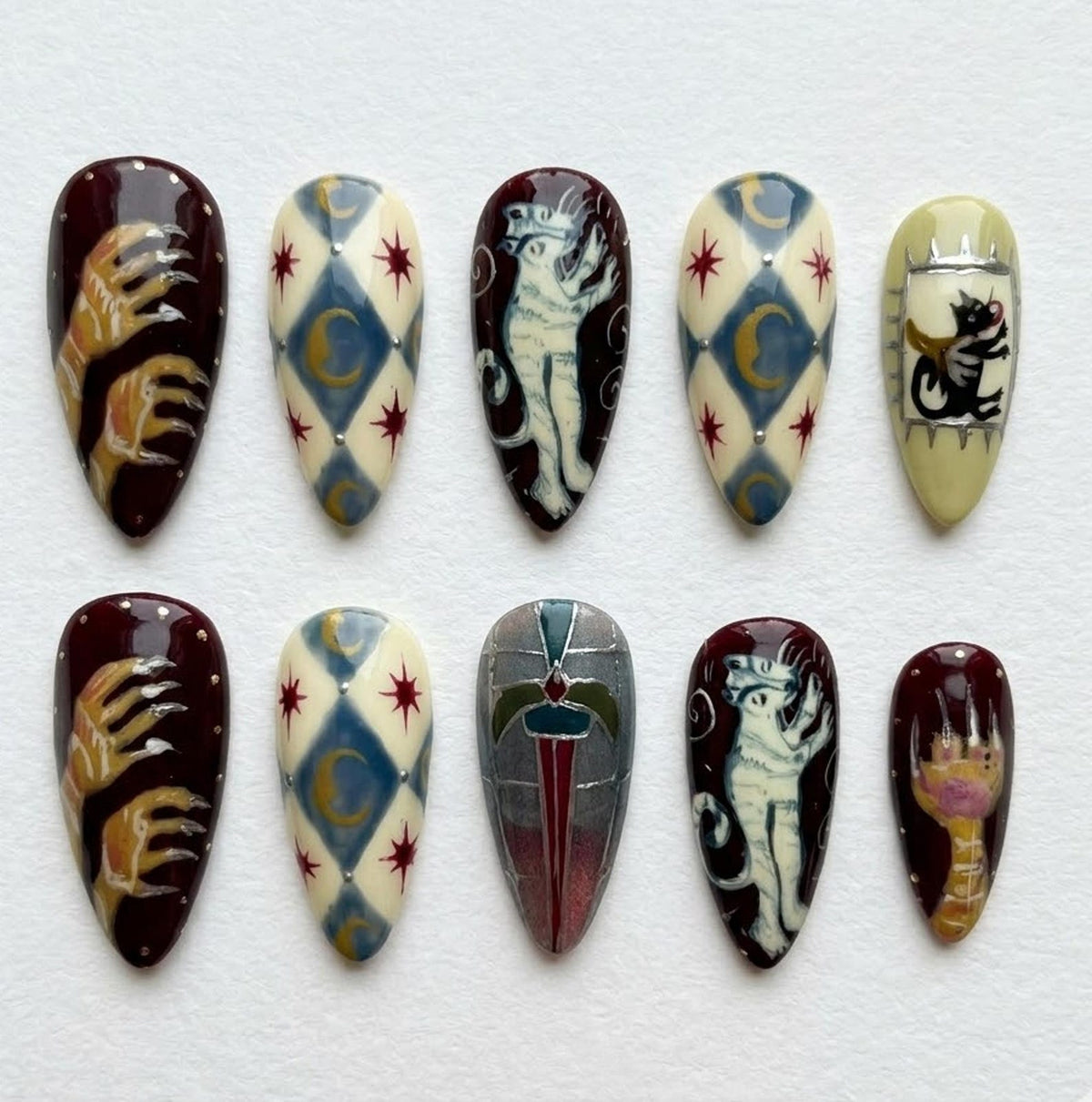 A set of ten almond-shaped false nails with intricate designs. The nails feature a variety of patterns, including a cat, a hand with claws, and geometric shapes in shades of burgundy, cream, blue, and gold.