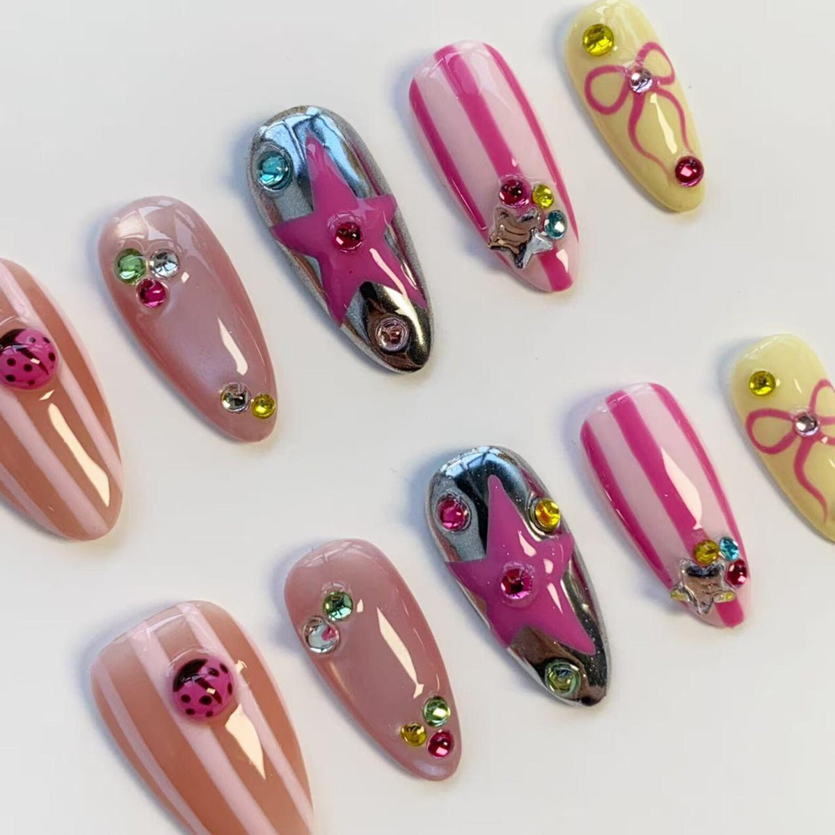 A collection of artificial nails in various designs. Some nails are pink with white stripes, others are a metallic silver with a pink star, and some are light pink with colorful rhinestones. A few nails feature a ladybug or a bow.