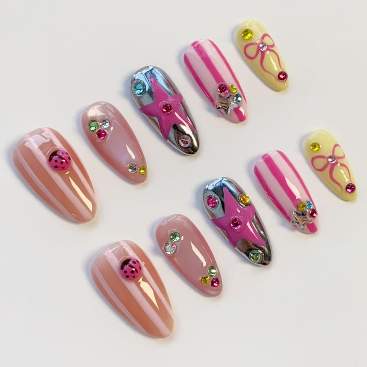 A collection of ten artificial nails in various designs. The nails feature pink, white, and silver accents, including stripes, stars, bows, and ladybug embellishments. Some nails are adorned with rhinestones, creating a playful and stylish aesthetic.