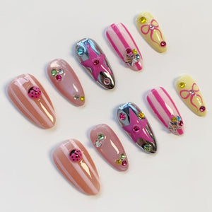 A collection of ten artificial nails in various designs. The nails feature pink, white, and silver accents, including stripes, stars, bows, and ladybug embellishments. Some nails are adorned with rhinestones, creating a playful and stylish aesthetic.