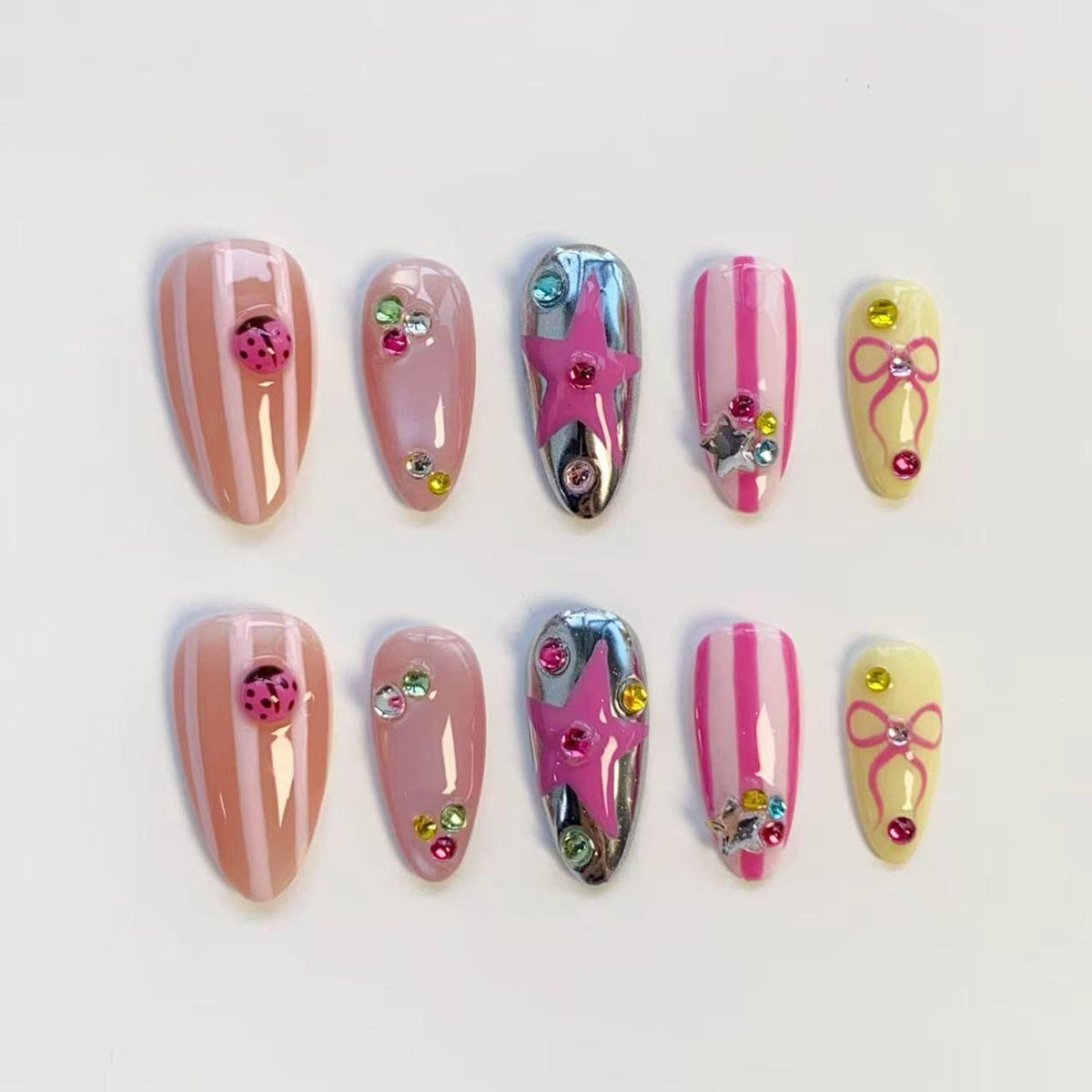 A set of ten artificial nails in various designs. The nails feature a mix of pink, yellow, and silver colors. Some nails have stripes, ladybug accents, or star and bow embellishments. Others are adorned with colorful rhinestones.