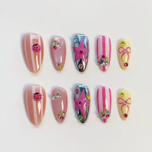 A set of ten artificial nails in various designs. The nails feature a mix of pink, yellow, and silver colors. Some nails have stripes, ladybug accents, or star and bow embellishments. Others are adorned with colorful rhinestones.