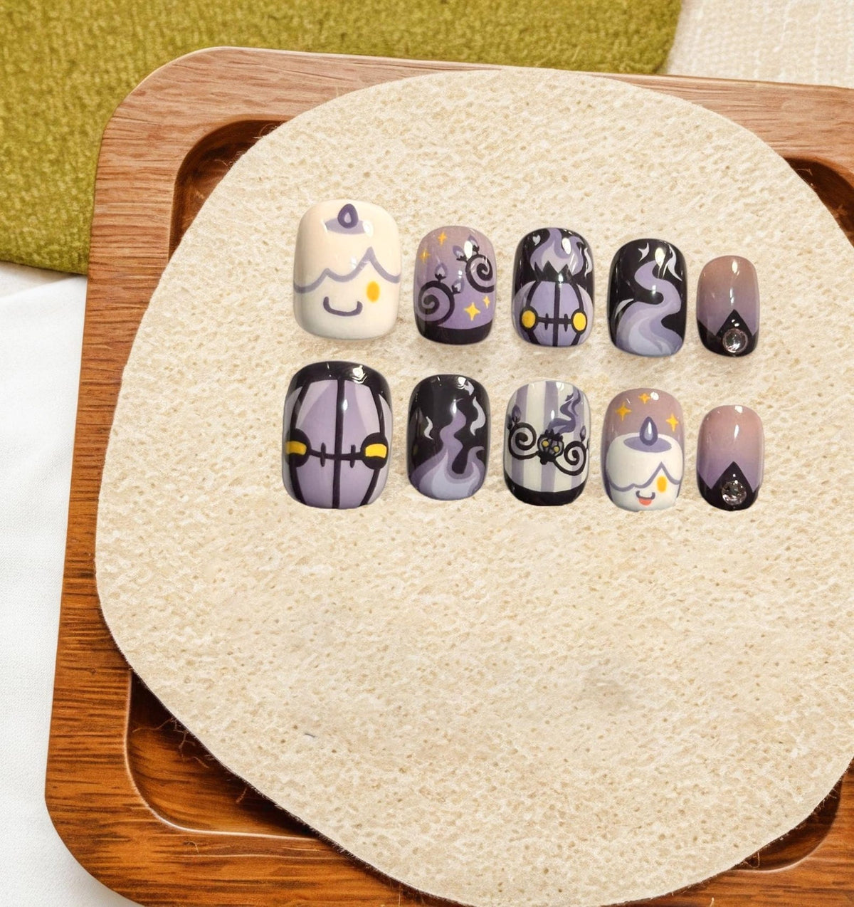 A set of ten press-on nails featuring a purple, black, and white color palette with designs including cartoon candles, swirling smoke patterns, striped motifs, and small clear rhinestones set against a neutral background.