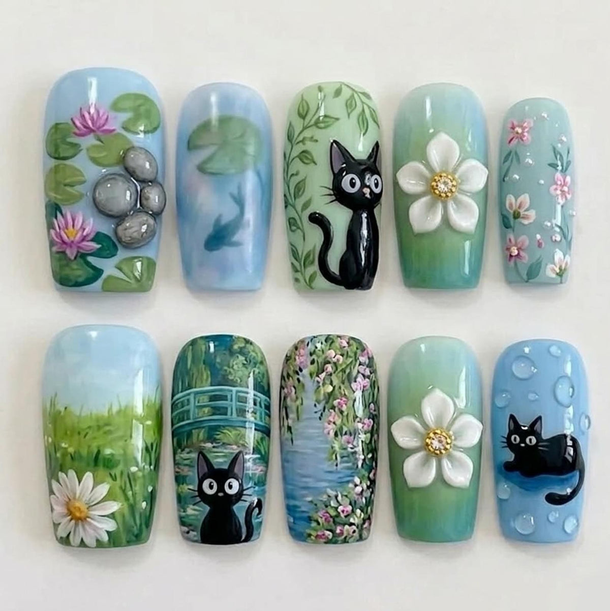 A set of ten artificial nails with various hand-painted designs. The designs include water lilies, a fish, a black cat, white flowers, and floral patterns. The nails are painted in shades of blue, green, and white.