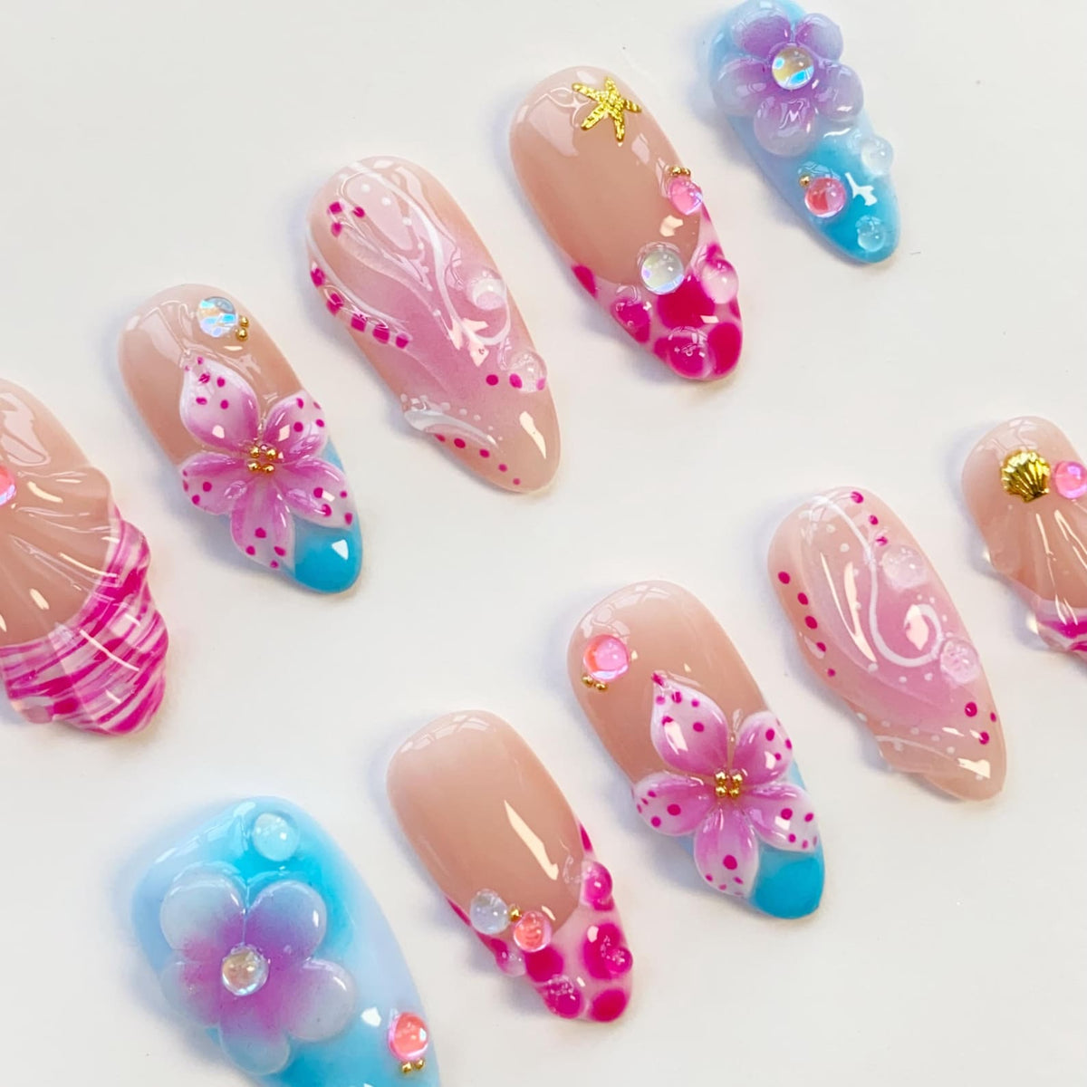 A collection of artificial nails with various designs. Some nails feature pink floral accents, while others have abstract patterns in pink and white. Several nails have a nude base with colorful embellishments and a turquoise or blue base.