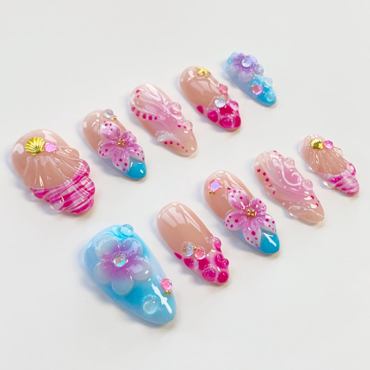 A collection of ten artificial nails with various designs. The nails feature a nude base with pink, blue, and white accents. Some nails have floral and seashell embellishments, while others have glitter and gem details. The nails are arranged on a wh