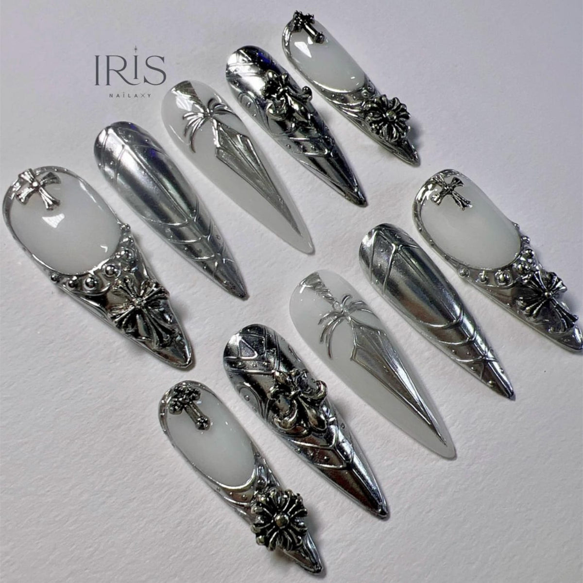 A collection of ten long, pointed artificial nails. The nails feature a combination of silver metallic and white designs, including crosses, floral accents, and intricate patterns. The brand name "IRIS NAILART" is visible in the upper left corner.