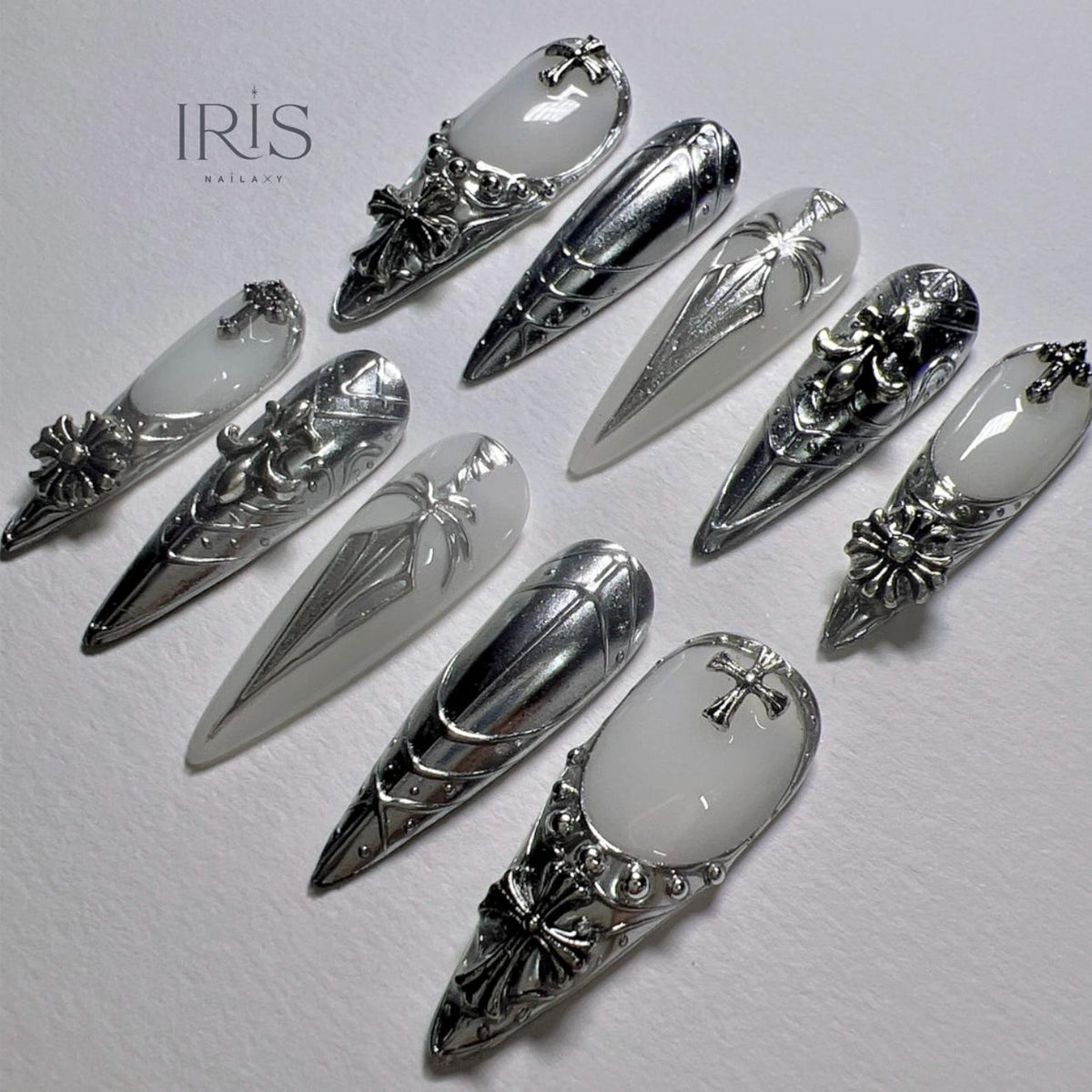 An assortment of silver nail art designs. Some nails feature a white base with silver embellishments, including crosses and floral patterns. The nails are long and pointed, with intricate metalwork.
