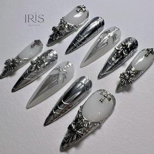 An assortment of silver nail art designs. Some nails feature a white base with silver embellishments, including crosses and floral patterns. The nails are long and pointed, with intricate metalwork.