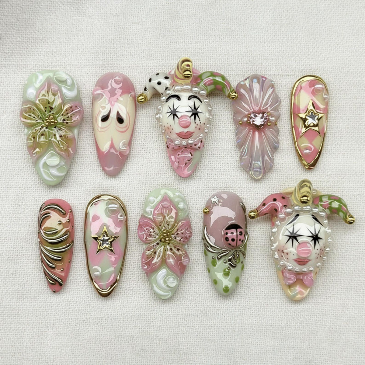 A collection of ten artificial nails with various designs. The nails feature a mix of colors, including pink, green, and gold. Some nails have floral designs, while others showcase a clown face or a star. The nails are arranged on a white surface.