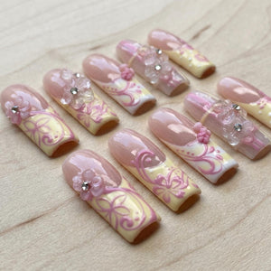 Set of ten artificial nails with a light pink and yellow floral design. Each nail features a clear top coat, with some adorned with small, decorative flowers and rhinestones. The nails are arranged on a light wood surface.