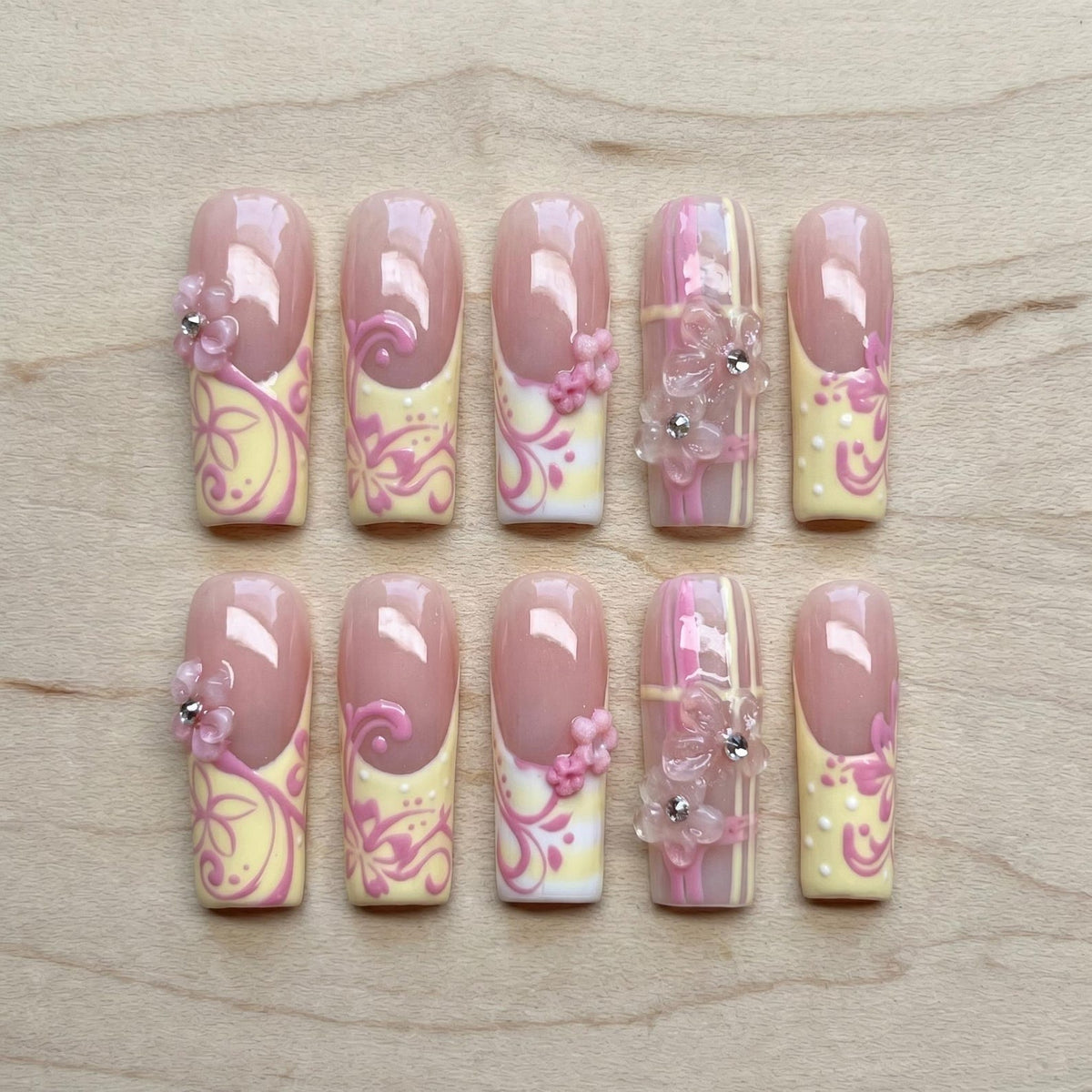 A set of ten artificial nails with a light pink base and a French tip design. The nails feature yellow and pink floral patterns, with some adorned with small flower embellishments and rhinestones. The nails are arranged on a wooden surface.