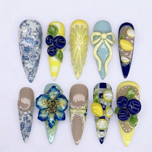 A collection of ten long, almond-shaped artificial nails. Each nail features unique designs, including floral patterns, fruit-inspired embellishments like lemons and blueberries, and bow accents. The color palette includes blues, yellows, and neutral