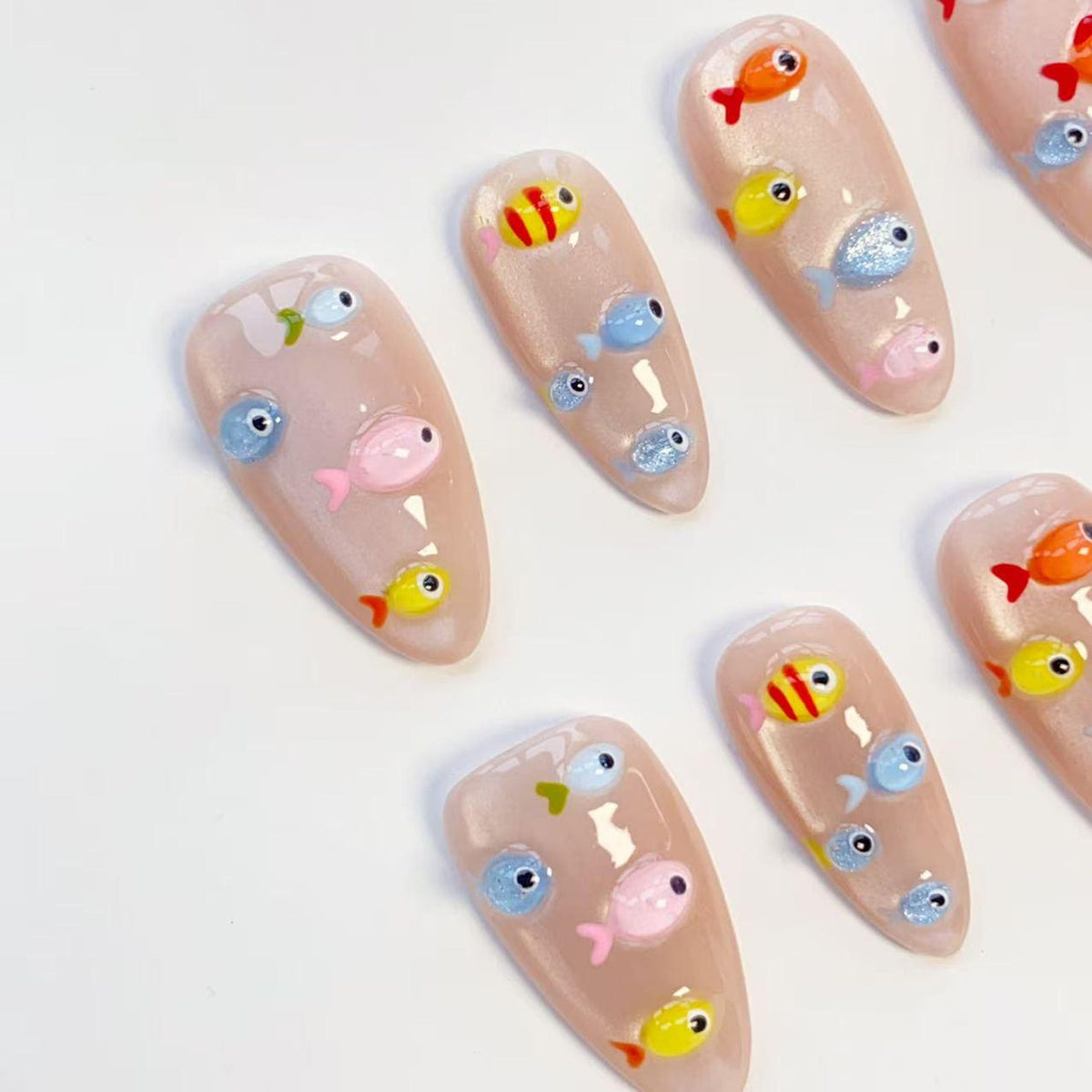 A set of almond-shaped artificial nails, each adorned with colorful, cartoonish fish designs. The fish are in shades of orange, yellow, pink, and blue, with some featuring glitter accents. The nails have a glossy, nude-toned base.
