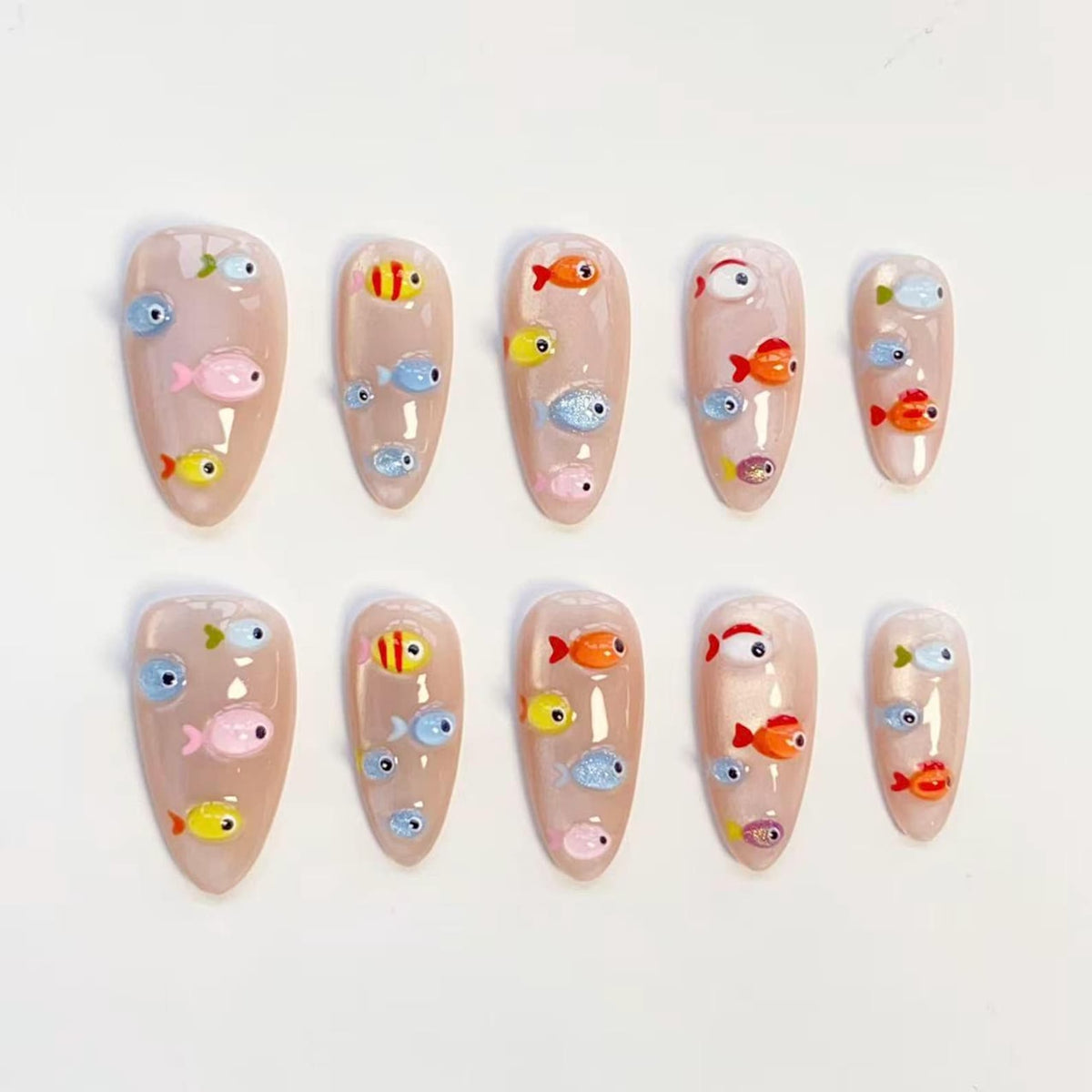 Set of ten artificial nails with a nude base, each adorned with colorful, cartoon fish designs. The fish are in shades of yellow, orange, pink, blue, and silver, creating a playful, aquatic theme. The nails are oval-shaped.