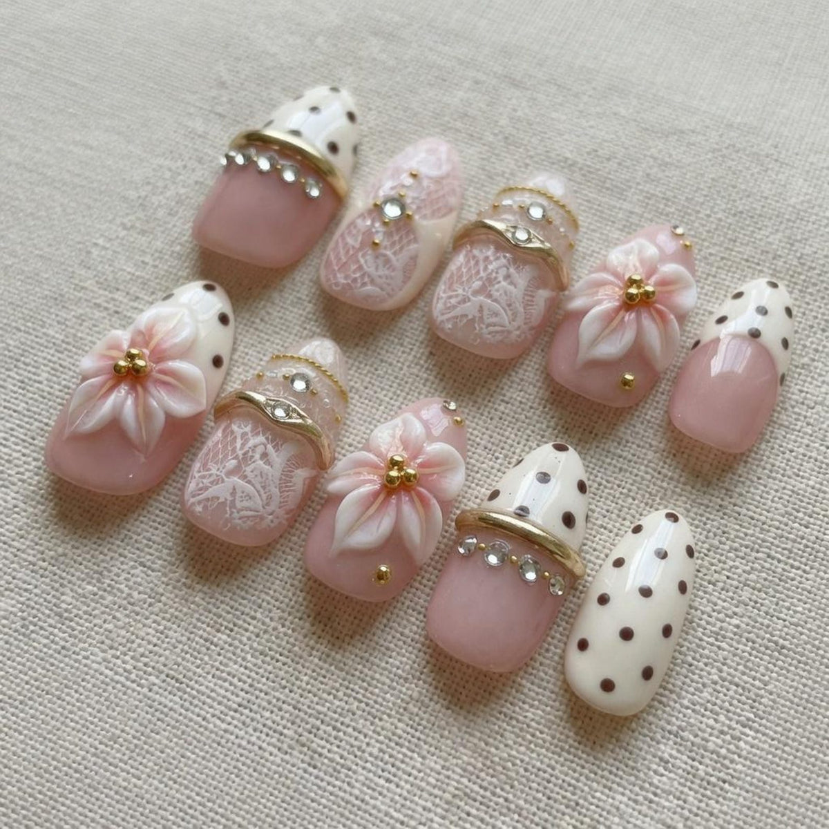 A set of ten press-on nails featuring a soft pink and cream color palette. The designs include raised 3D floral accents, white lace patterns, brown polka dots, and gold-toned metallic bands embellished with small clear rhinestones.