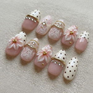 A set of ten press-on nails featuring a soft pink and cream color palette. The designs include raised 3D floral accents, white lace patterns, brown polka dots, and gold-toned metallic bands embellished with small clear rhinestones.