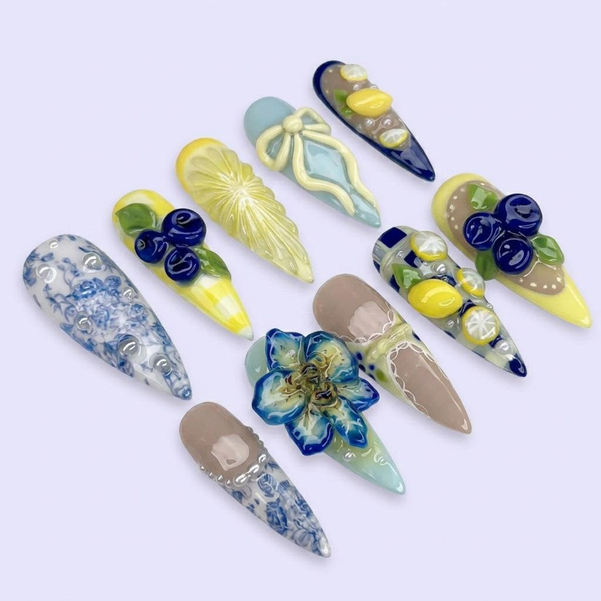 A collection of ten long, almond-shaped artificial nails. Each nail features unique designs, including floral patterns, fruit, and bow accents. Colors include blue, yellow, brown, and white. The nails are arranged on a light purple surface.