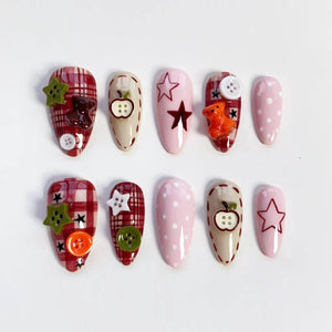 A set of ten artificial nails in various designs. Some nails feature a red plaid pattern, while others are solid pink or beige. Decorations include stars, buttons, and small teddy bears. The nails are oval-shaped.