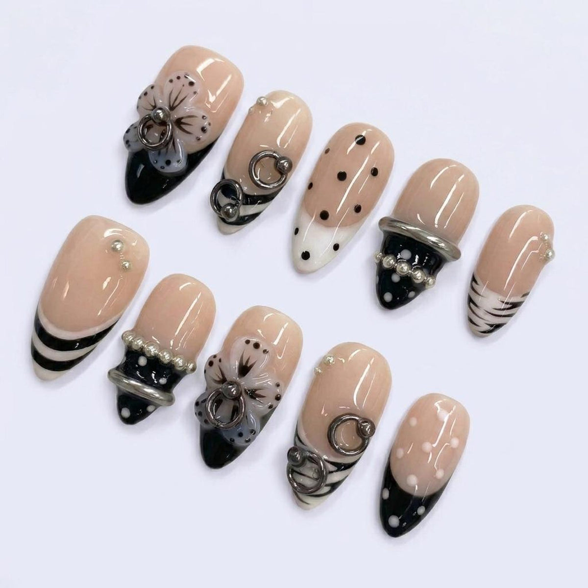 A set of ten artificial nails in various designs. The nails feature a nude base with black and white accents, including stripes, polka dots, and floral embellishments. Some nails have silver rings and pearl accents.