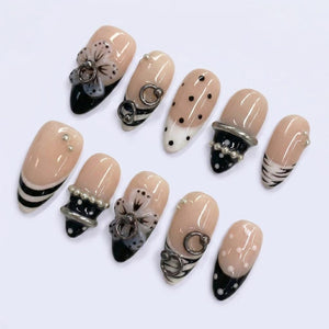 A set of ten artificial nails in various designs. The nails feature a nude base with black and white accents, including stripes, polka dots, and floral embellishments. Some nails have silver rings and pearl accents.