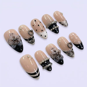 A set of ten almond-shaped artificial nails. Each nail features a nude base with various designs, including black and white accents, polka dots, floral embellishments, and silver rings. Some nails have pearl accents.