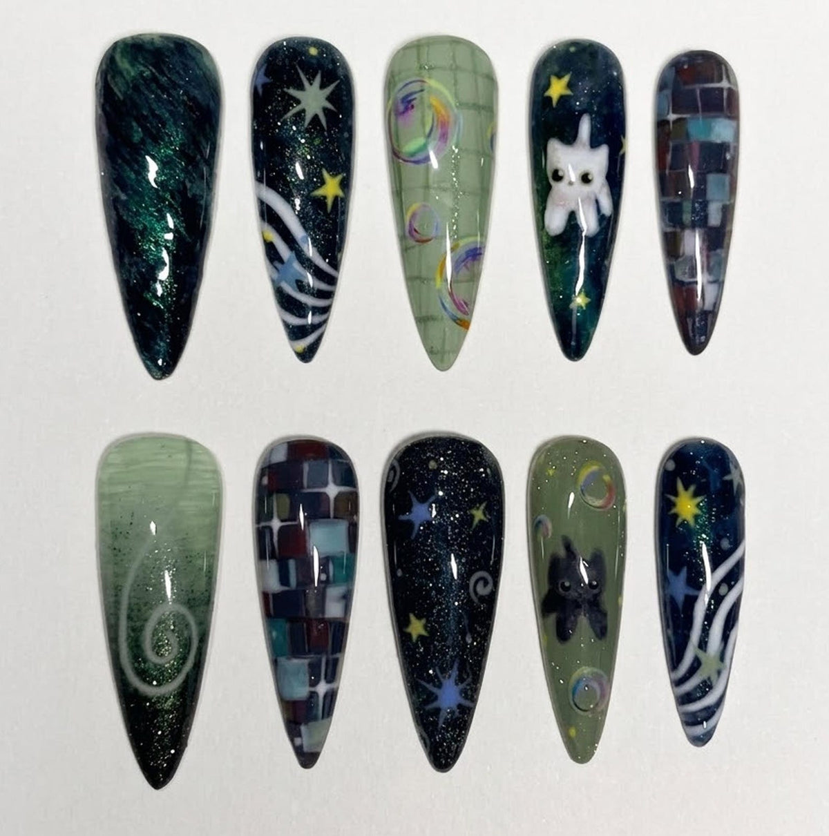 A set of ten almond-shaped press-on nails featuring various designs including dark green marbled patterns, deep blue starry night skies with white cat illustrations, light green bubble motifs, and dark multi-colored mosaic tile patterns.
