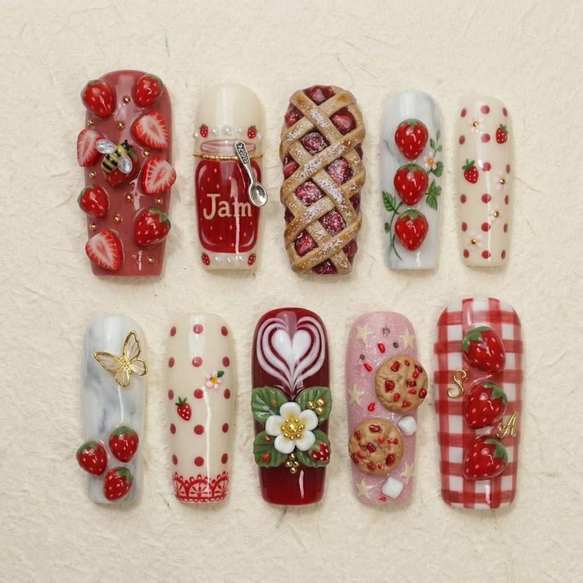 A set of ten press-on nails featuring intricate 3D designs including strawberries, a jam jar with a spoon, a lattice pie, a butterfly, cookies, and floral patterns. The nails display various textures, red and white colors, and gold accents.