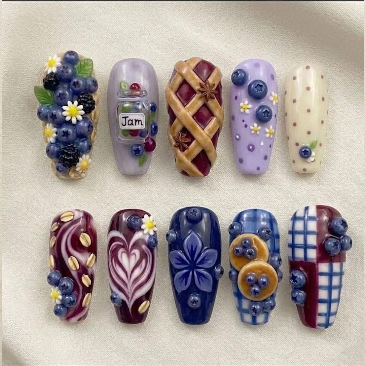 A set of ten press-on nails featuring 3D designs including blueberries, blackberries, daisies, a jar labeled Jam, a lattice pie crust, polka dots, oat-like shapes, heart latte art, a blue flower, pancakes, and blue plaid patterns.