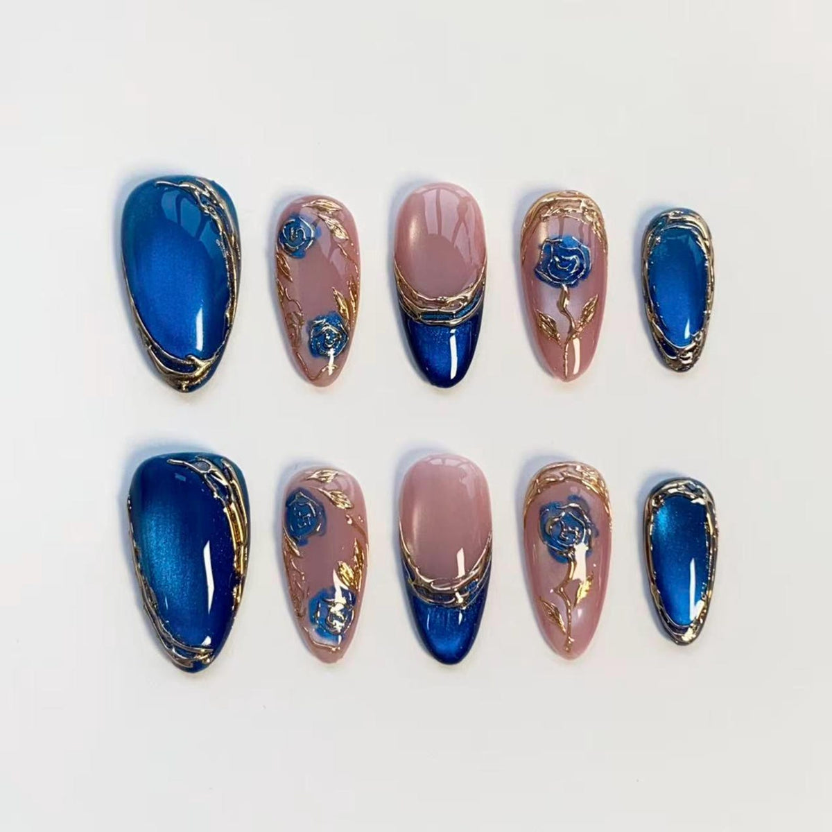 Set of ten oval-shaped false nails. The nails feature a variety of designs, including solid blue with gold trim, pink with blue rose accents, and pink with a blue tip and gold trim. The nails are arranged on a white surface.