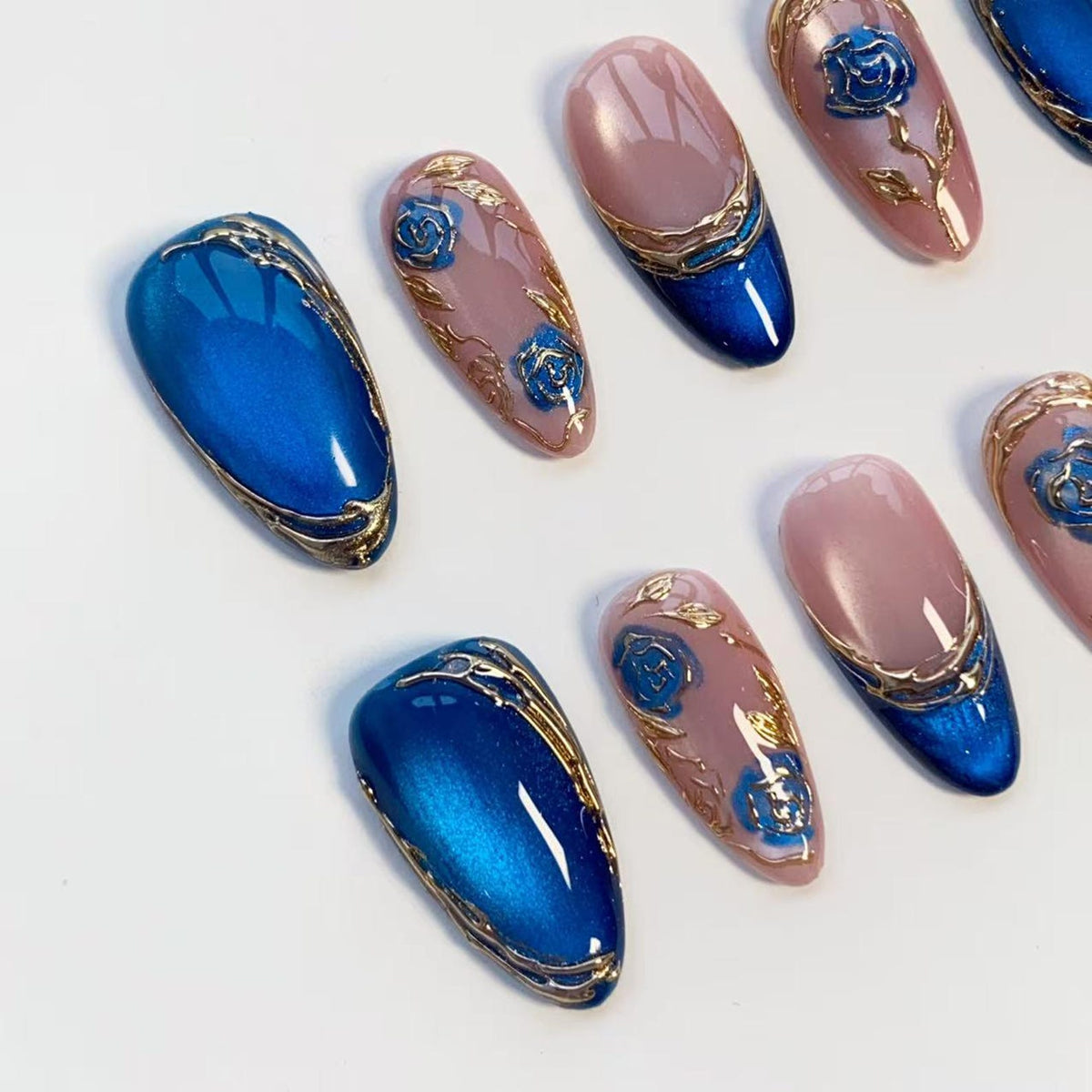 A collection of artificial nails with intricate designs. The nails feature a combination of blue, pink, and gold accents. Some nails have a solid blue color, while others showcase floral patterns and gold embellishments. The overall aesthetic is eleg