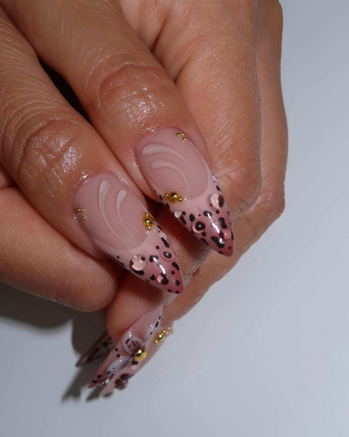 Long, almond-shaped nails with a nude base, adorned with a swirl design and dark brown leopard print tips. Gold embellishments add a touch of sparkle. The nails are glossy and well-manicured.