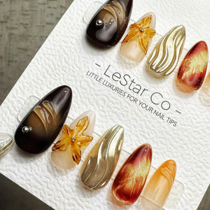 A set of almond-shaped press-on nails on a white display card labeled LeStar Co. LITTLE LUXURIES FOR YOUR NAIL TIPS. The nails feature various designs including dark brown, gold metallic, amber floral