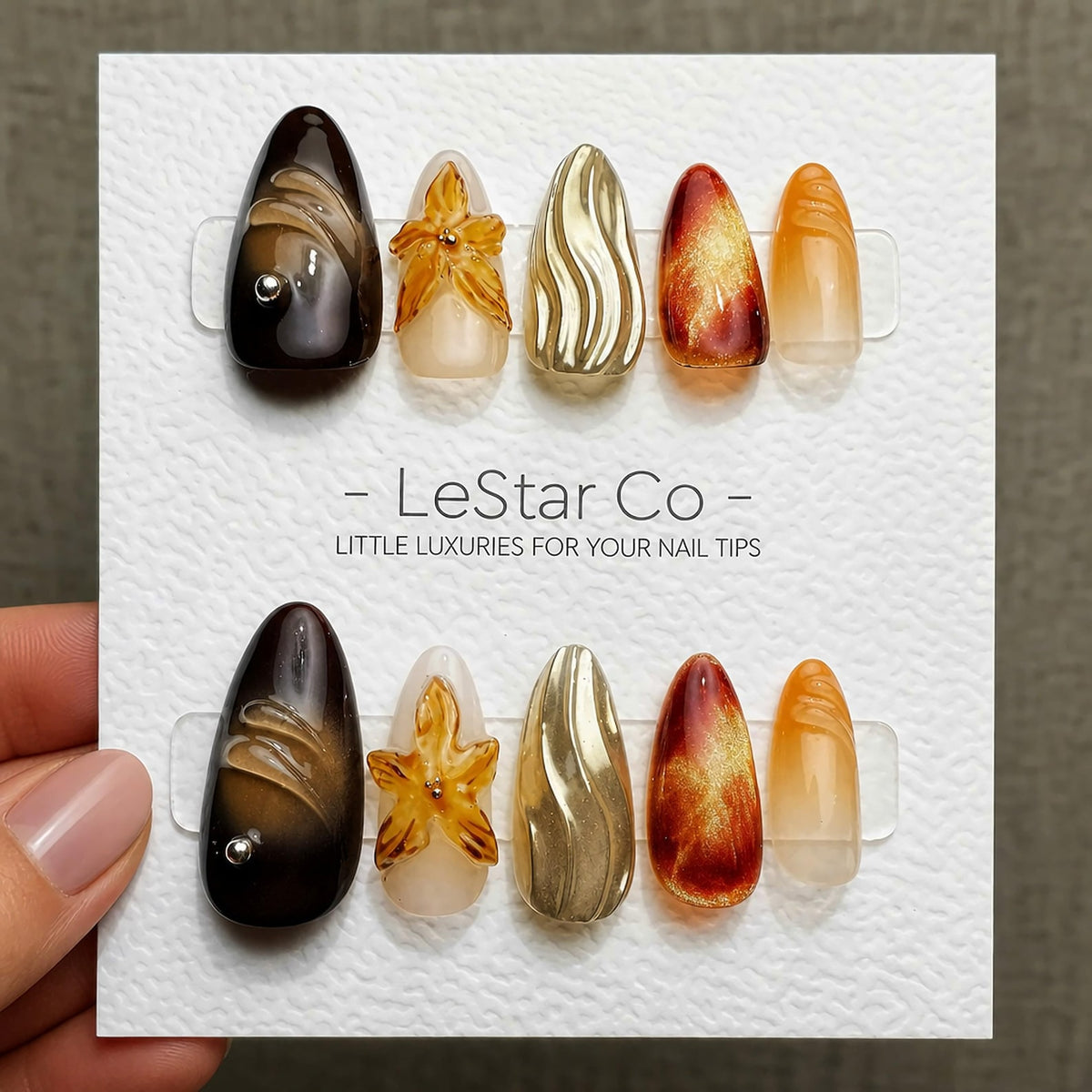 A set of ten press-on nails arranged on a white display card labeled LeStar Co - LITTLE LUXURIES FOR YOUR NAIL TIPS. The nails feature almond shapes with varied designs including dark brown, amber flo