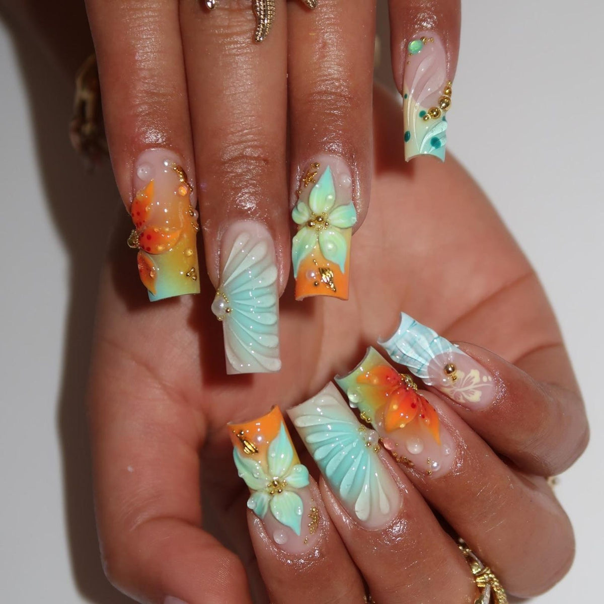 Long square-shaped artificial nails featuring a gradient of orange and light blue. The nails are decorated with three-dimensional sculpted flower petals, raised fan-like patterns, small gold metallic beads, and clear dew-like accents.