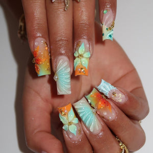 Long square-shaped artificial nails featuring a gradient of orange and light blue. The nails are decorated with three-dimensional sculpted flower petals, raised fan-like patterns, small gold metallic beads, and clear dew-like accents.