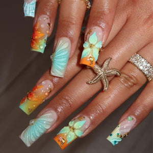 Long square-shaped artificial nails featuring a gradient of orange and light blue. The designs include three-dimensional sculpted flowers, shell patterns, small gold beads, and tiny white pearls. A gold starfish ring is worn on one finger.