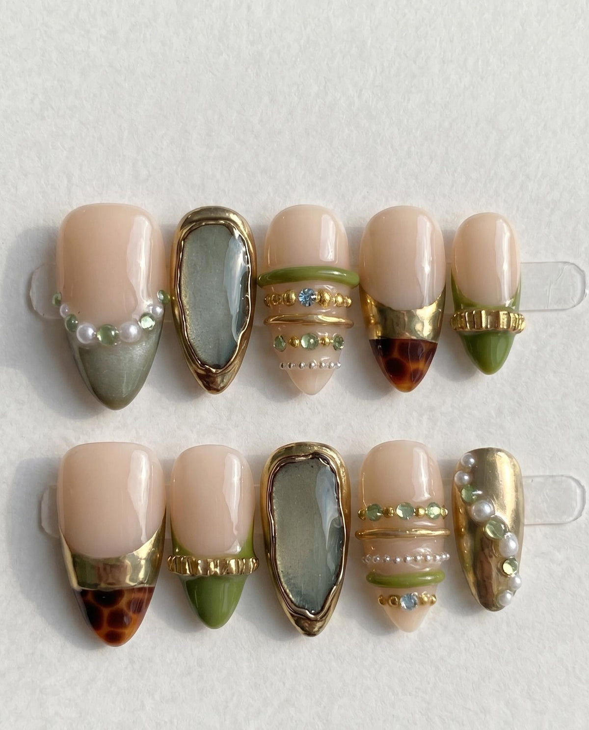 A set of ten press-on nails featuring a nude base with olive green tips, gold metallic accents, and small embedded pearls and crystals. The designs include tortoiseshell patterns, textured gold bands, and smooth, glossy finishes.
