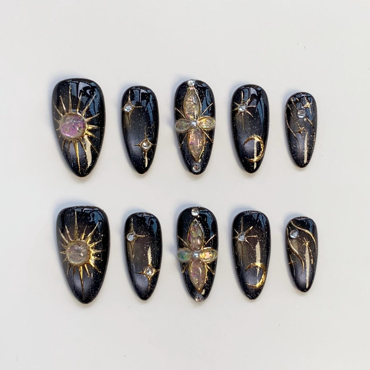 A set of ten black and gold press-on nails. Each nail features celestial designs, including suns, stars, and crescent moons, with iridescent and rhinestone accents. The nails are oval-shaped and have a glossy finish.