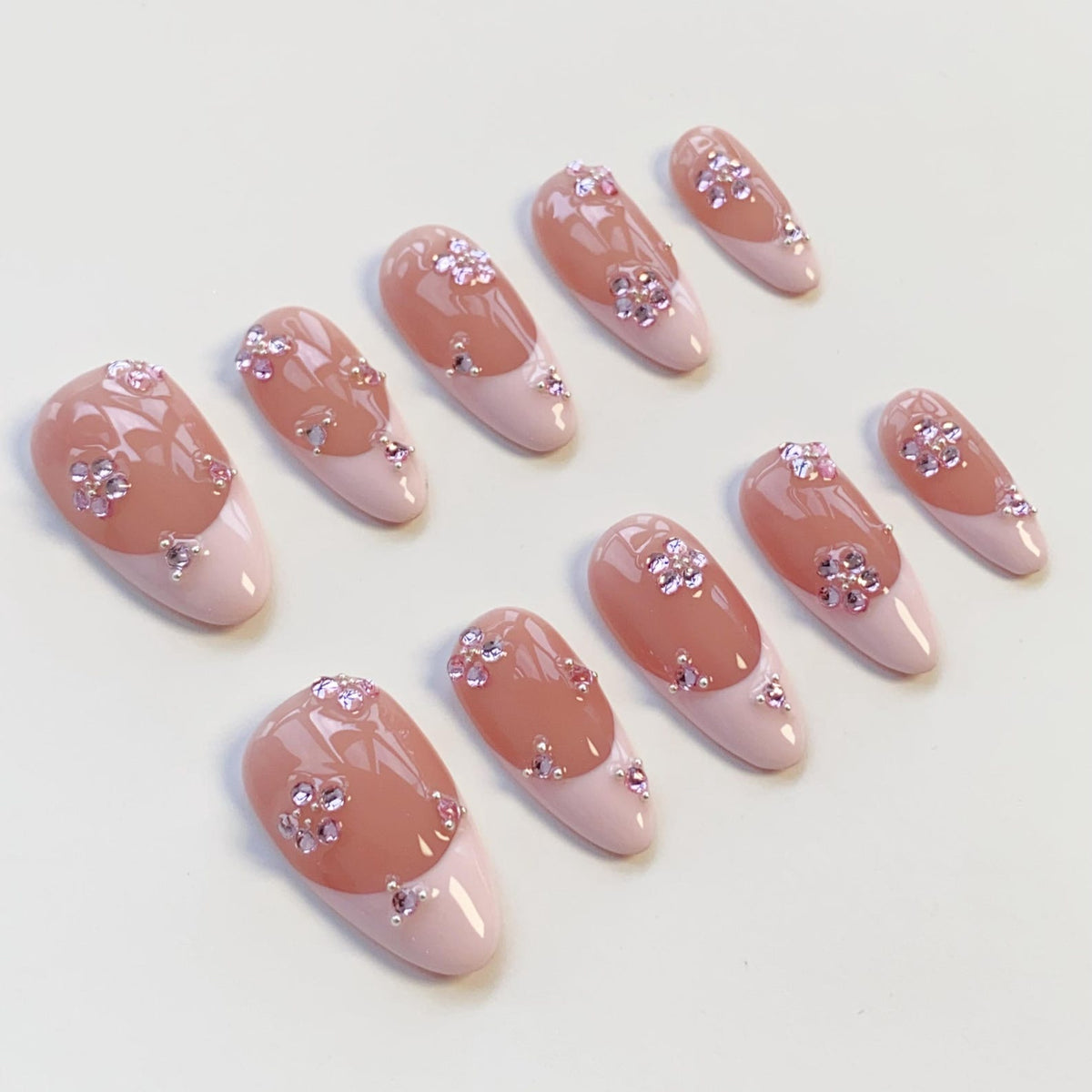 Set of ten artificial nails with a light pink base and a peach-colored top. Each nail features a floral design made of small, sparkling rhinestones. The nails have a rounded shape.